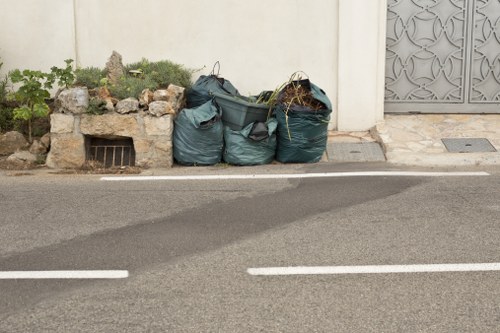 The image shows three large green refuse bags filled with household waste, positioned on the edge of a paved street next to a white wall. The bags are made of plastic with a slightly glossy finish and appear to contain mixed rubbish, possibly including garden waste and general household debris. They are placed on the roadside, directly adjacent to a painted white line separating the main road from the parking or loading area. Behind the bags, there is a small raised flower bed constructed from rocks and filled with green plants and small shrubs, providing a contrast to the urban environment. To the right, part of a decorative metal gate or door is visible, featuring a detailed geometric pattern. The scene is well-lit with natural daylight, emphasizing the textures of the bag material, the pavement, and the surrounding elements. This visual matches the context of rubbish removal services offered by Ruislip House Clearance, serving the local area around Ruislip, possibly within the HA postcode district, with an emphasis on waste disposal and clearance in residential streets.