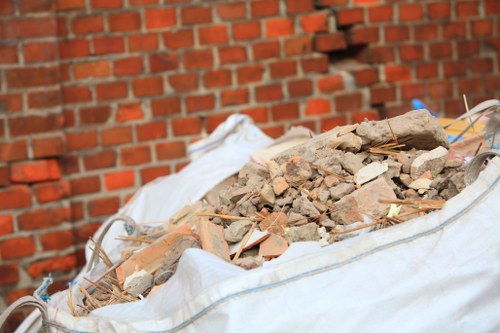 A large white rubble sack filled with a mixture of broken bricks, concrete pieces, and assorted debris sits on a paved surface outdoors against a red brick wall. The debris appears to be construction or demolition waste, with rough, uneven textures and varying shades of beige, brown, and reddish tones. The bag's fabric is slightly wrinkled and tied at the top, with some loose strands of wire and small fragments visible among the rubble. The scene is set in a residential or commercial outdoor area, possibly a driveway or pavement, where Ruislip House Clearance services might operate to remove waste from property renovations or clearance projects, located within the Ruislip region, close to postcode HA4 or HA5. The overall lighting is natural, suggesting daytime conditions, emphasizing the rough surface of the broken brick and concrete materials against the brick wall backdrop.