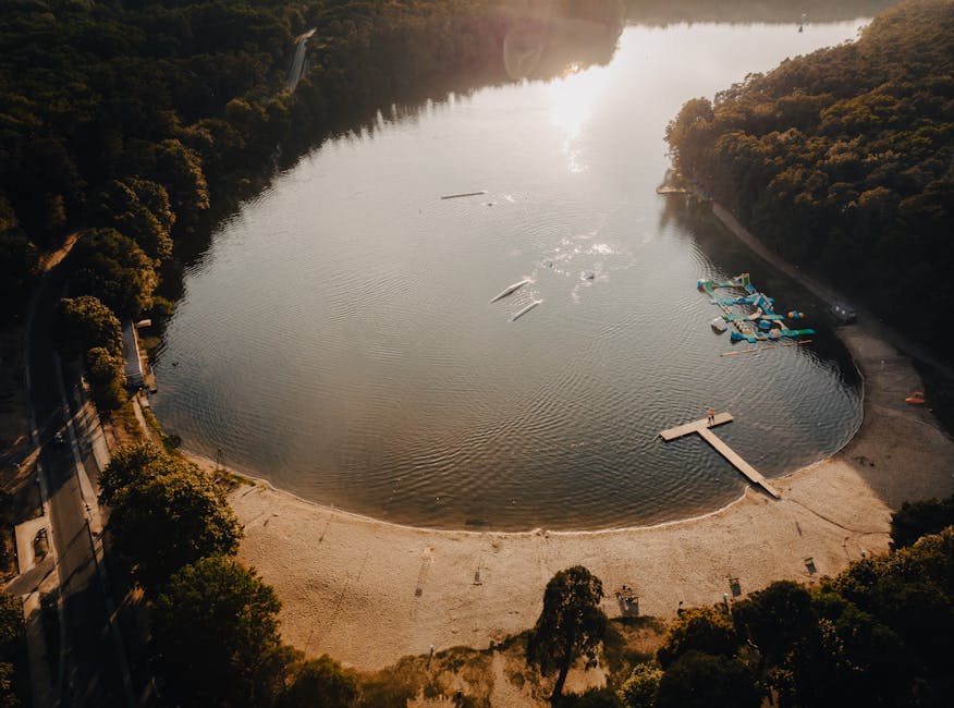 An aerial view of a large, freshwater reservoir surrounded by dense, forested hills. The reservoir features a curved sandy shoreline with a small, rectangular wooden pier extending into the water. Sev