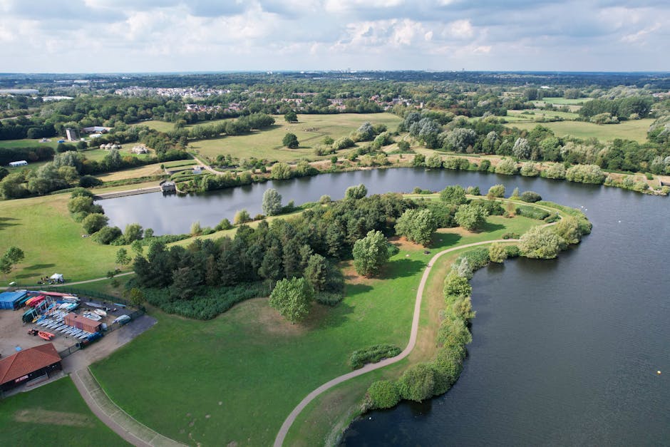 An aerial view of a large, freshwater reservoir surrounded by dense, forested hills. The reservoir features a curved sandy shoreline with a small, rectangular wooden pier extending into the water. Sev
